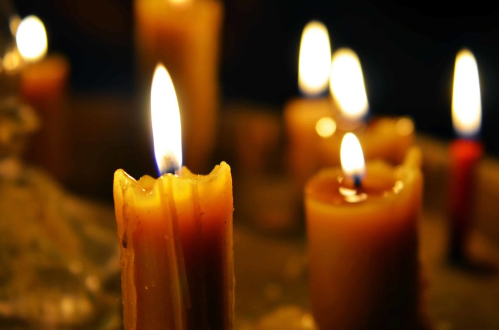 A close-up of several lit candles in a dimly lit setting. The candles are partially melted, with warm flames flickering gently, creating a soft, glowing ambiance. The background is dark, enhancing the brightness of the candlelight.