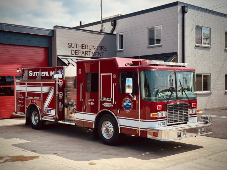 A red fire truck labeled "Sutherlin Fire Department" is parked outside a modern fire station with red garage doors and gray walls. The truck has white striping and various emergency equipment.