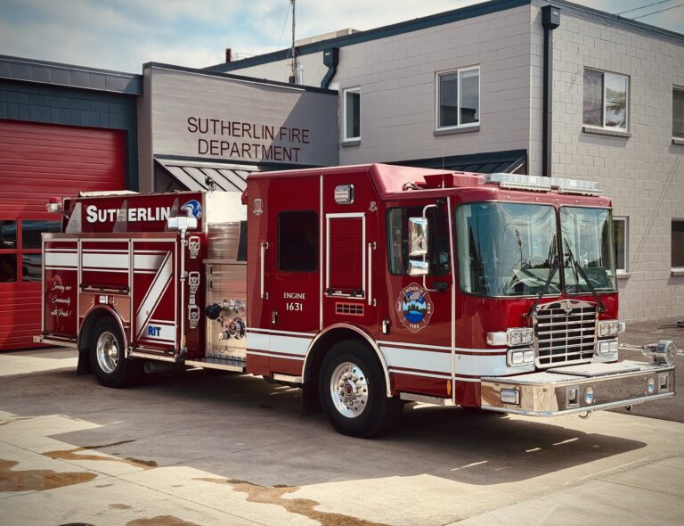 A red Sutherlin Fire Department fire engine is parked outside a fire station with red garage doors and a gray building. The truck displays department insignia and reflective stripes.