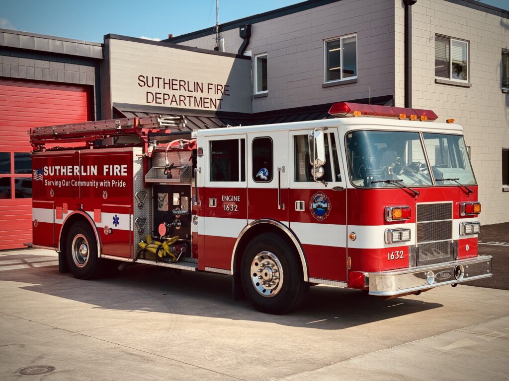 A red and white fire truck labeled "Sutherlin Fire" is parked outside the Sutherlin Fire Department building on a sunny day. The fire truck has lights on top and equipment visible on the sides.