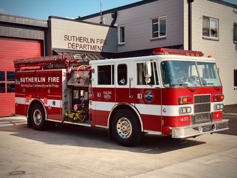 A red and white fire truck labeled "Sutherlin Fire" is parked outside the Sutherlin Fire Department building on a sunny day. The fire truck has lights on top and equipment visible on the sides.
