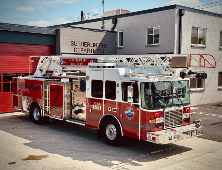 A red and white fire truck labeled "Truck 1651" is parked outside the Sutherlin Fire Department building with a red garage door in the background.