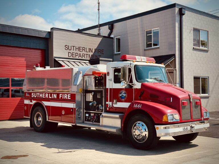 A red and white fire truck labeled "Sutherlin Fire" is parked outside the Sutherlin Fire Department building on a sunny day. The truck has emergency lights, equipment compartments, and the number 1661 on its bumper.
