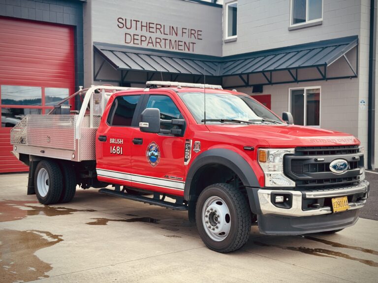 A red brush fire truck labeled "BRUSH 1681" is parked outside the Sutherlin Fire Department building, with the department name visible above the entrance.
