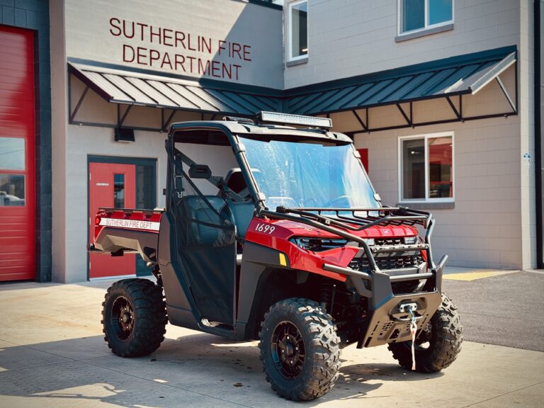 A red and black off-road utility vehicle marked "Sutherlin Fire Dept" is parked outside the Sutherlin Fire Department building on a sunny day.