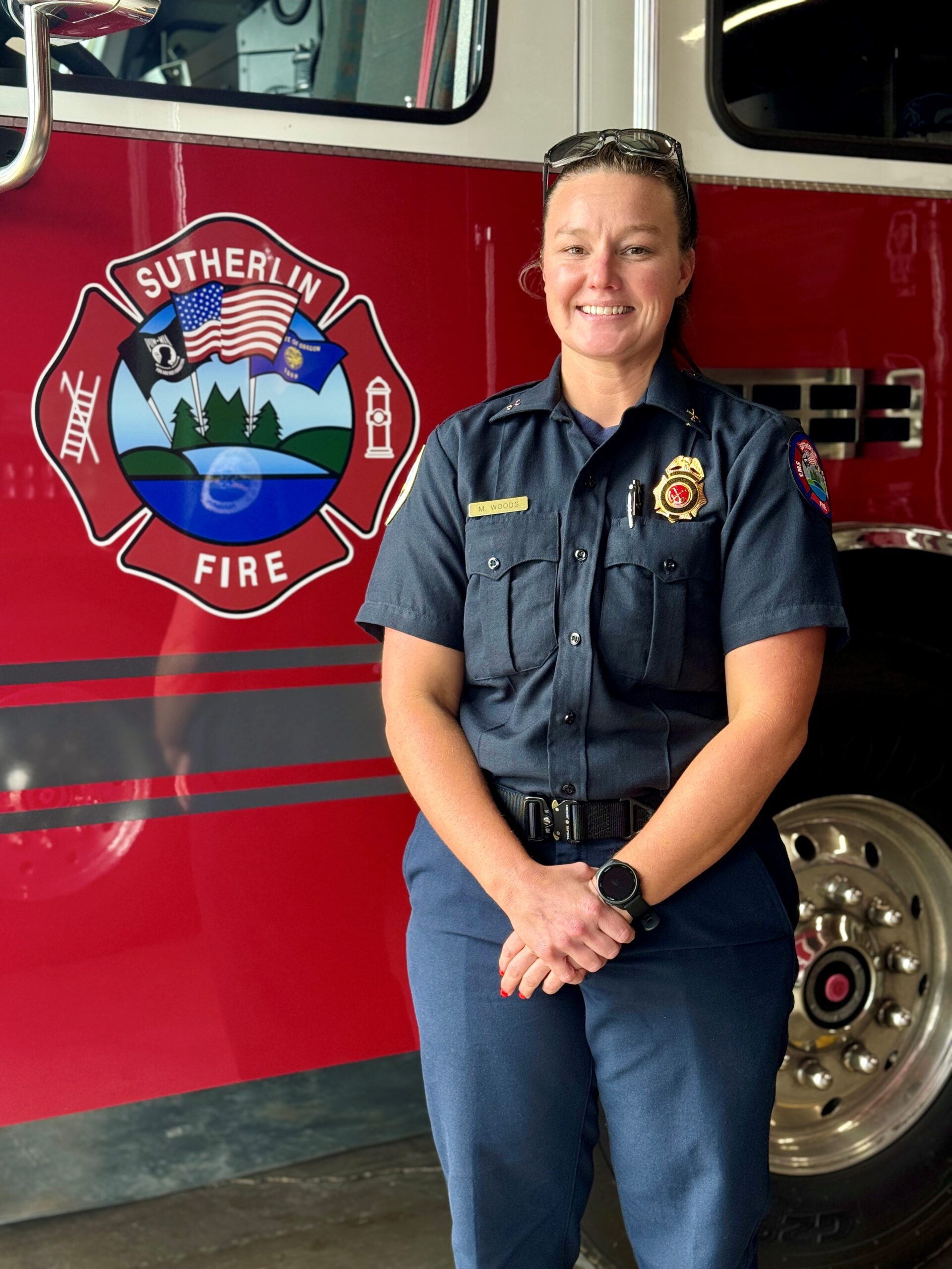 A firefighter in uniform stands smiling in front of a red fire truck with the "Sutherlin Fire" emblem visible on the side.