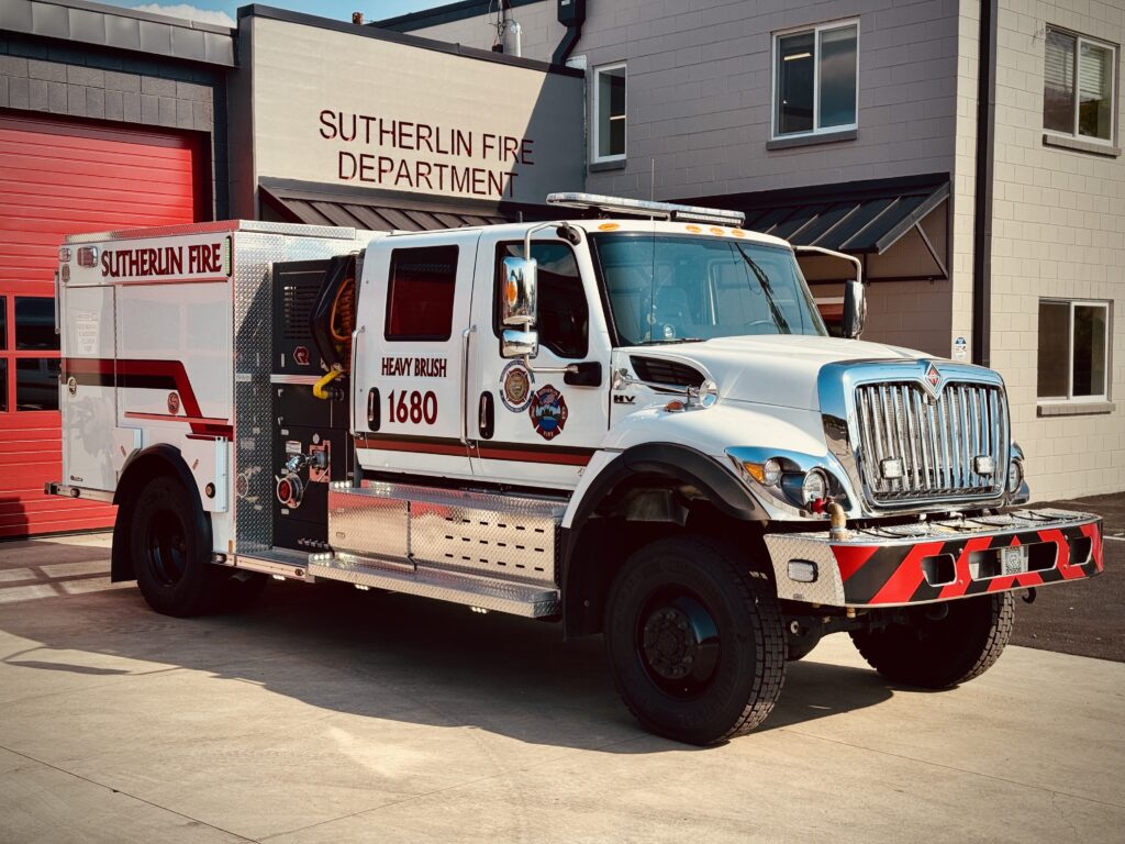 A white and red fire truck labeled "Sutherlin Fire Department" and "Heavy Brush 1680" is parked outside a fire station with a red garage door and beige walls.