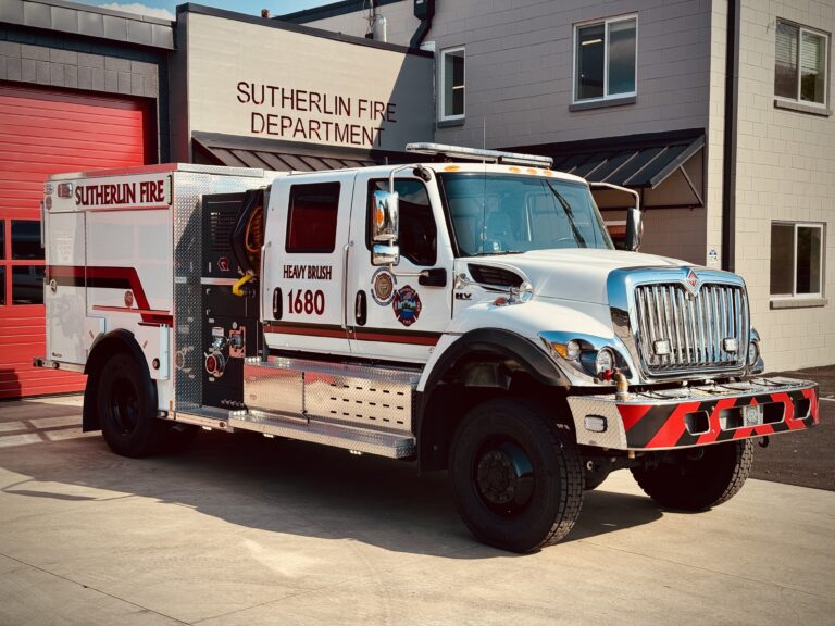 A white and red fire truck labeled "Sutherlin Fire Department" and "Heavy Brush 1680" is parked outside a fire station with a red garage door and beige walls.